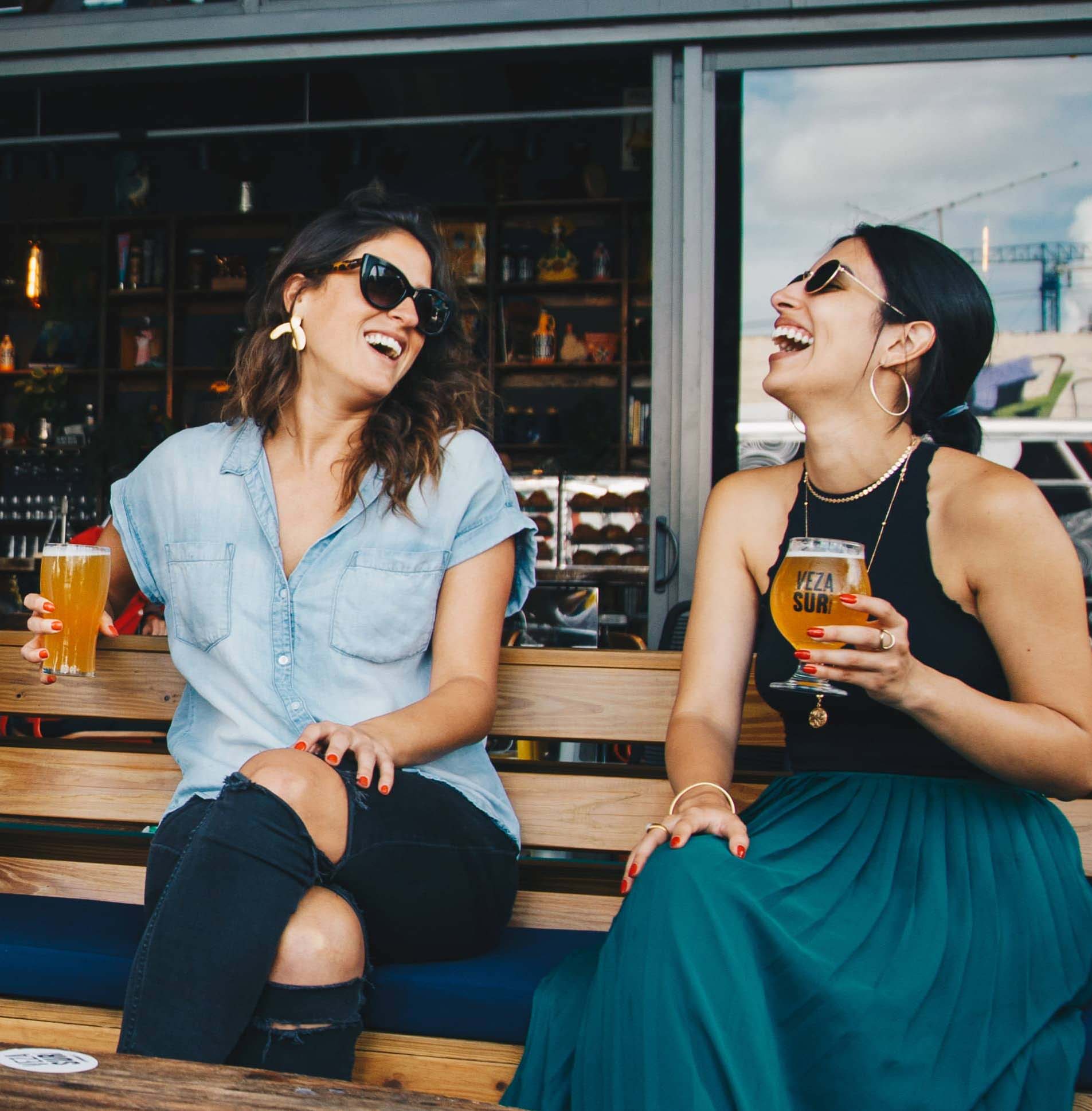 two women laughing while holding ciders at a bar